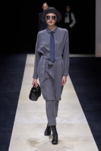 Model walks a fashion runway wearing a gray matching shirt-dress with a tied belt, navy patterned tie, black ankle boots, wide-brim hat, and round sunglasses, carrying a black textured handbag.