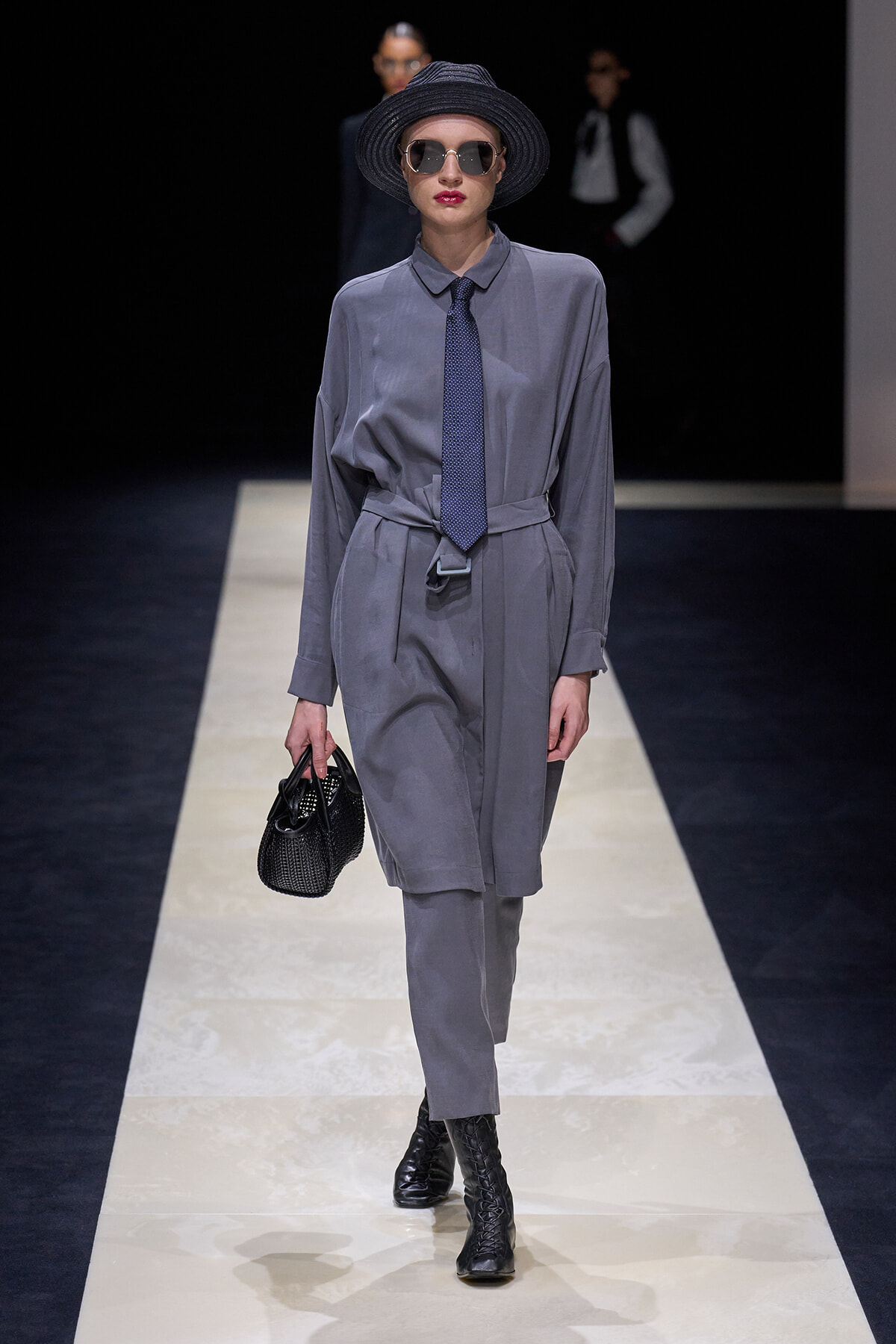 Model walks a fashion runway wearing a gray matching shirt-dress with a tied belt, navy patterned tie, black ankle boots, wide-brim hat, and round sunglasses, carrying a black textured handbag.