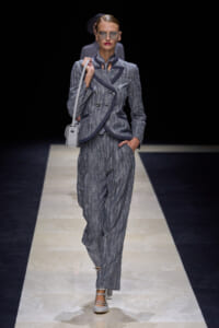Model walking a runway in a gray pinstripe pantsuit with navy trim, carrying a light gray shoulder bag.