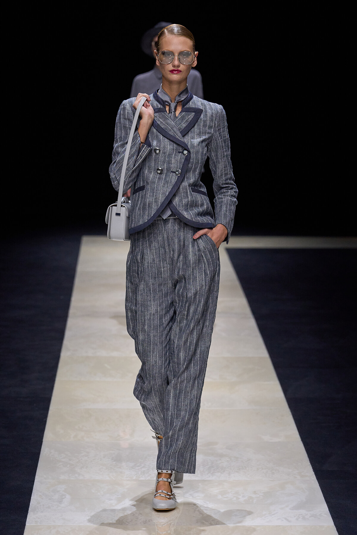 Model walking a runway in a gray pinstripe pantsuit with navy trim, carrying a light gray shoulder bag.