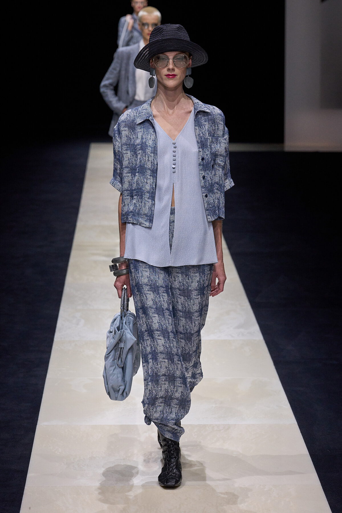 Model walks the runway in a light blue sleeveless blouse with matching patterned jacket and wide-leg pants, accessorized with a black hat, oversized glasses, bold earrings, and a light blue handbag.