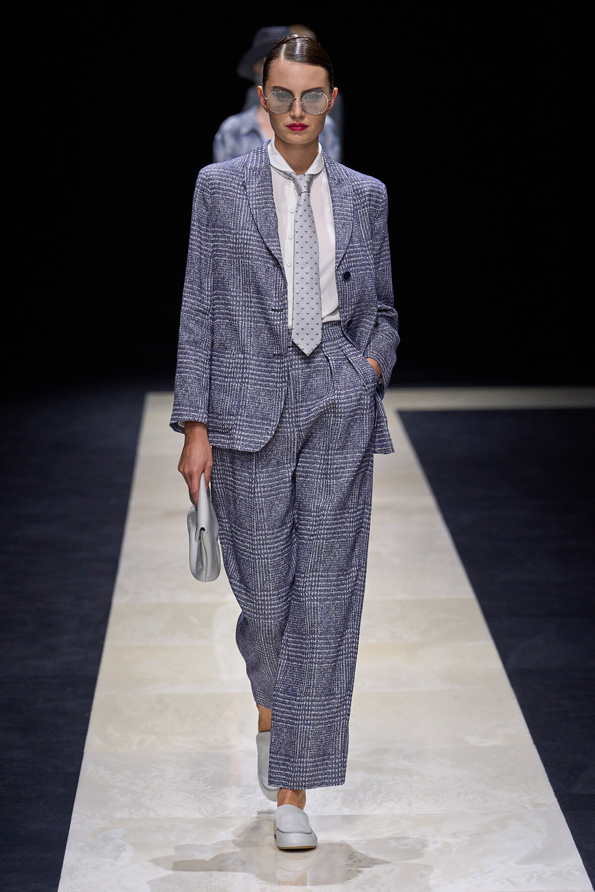 Female model on a runway wearing a blue-gray checkered suit with a matching blazer and wide-leg trousers, white shirt, light gray tie, silver glasses, and a small gray clutch.