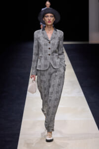 Model walking a fashion runway in a grey plaid suit with a black wide-brim hat, carrying a small light handbag.