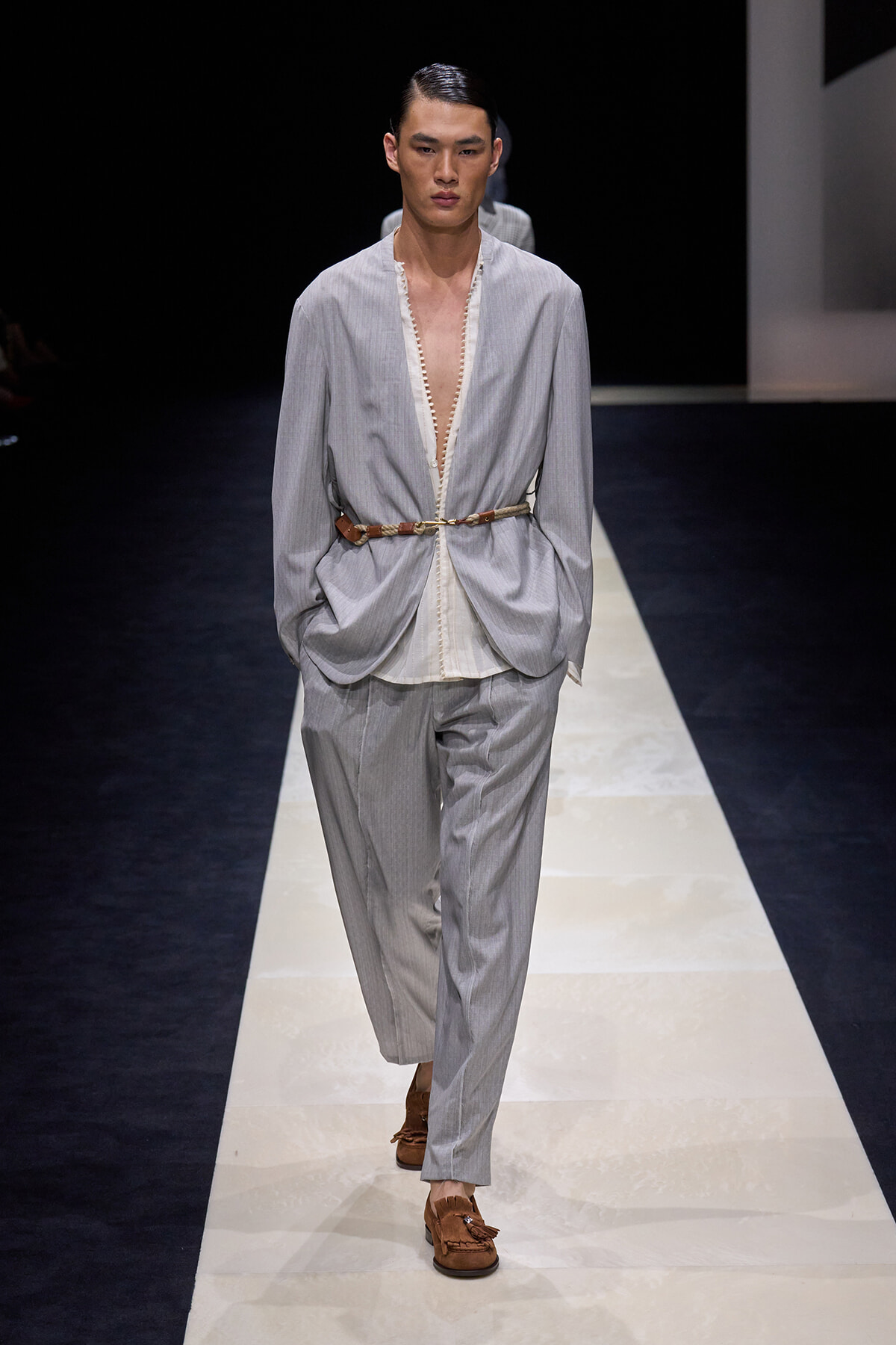 Male model on a runway wearing a light gray, loose suit with an open front shirt and a brown belt, brown loafers.