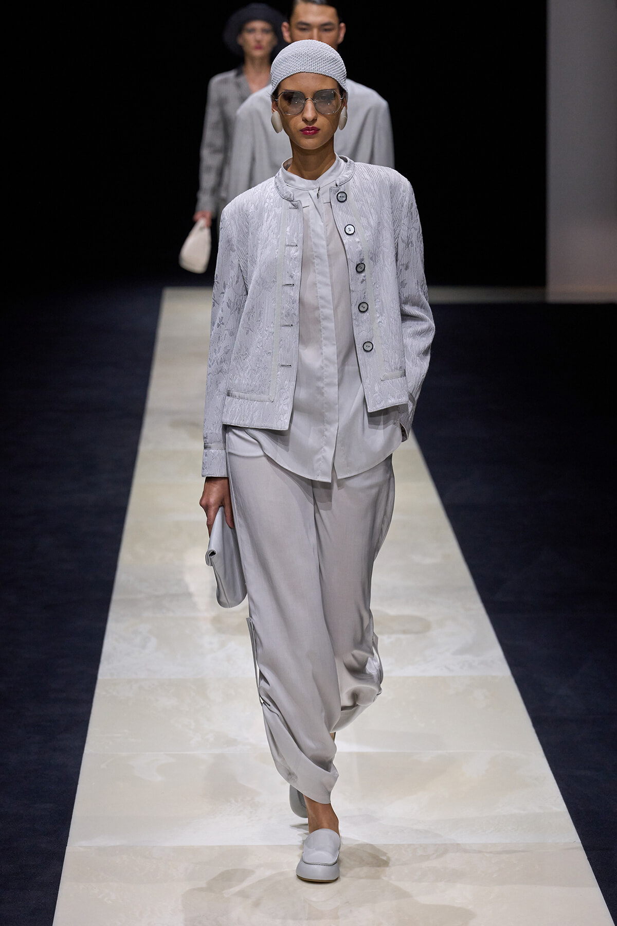 Model on a runway wearing a pale gray textured jacket with a matching blouse and loose trousers, accessorized with a headscarf, sunglasses, and a clutch.