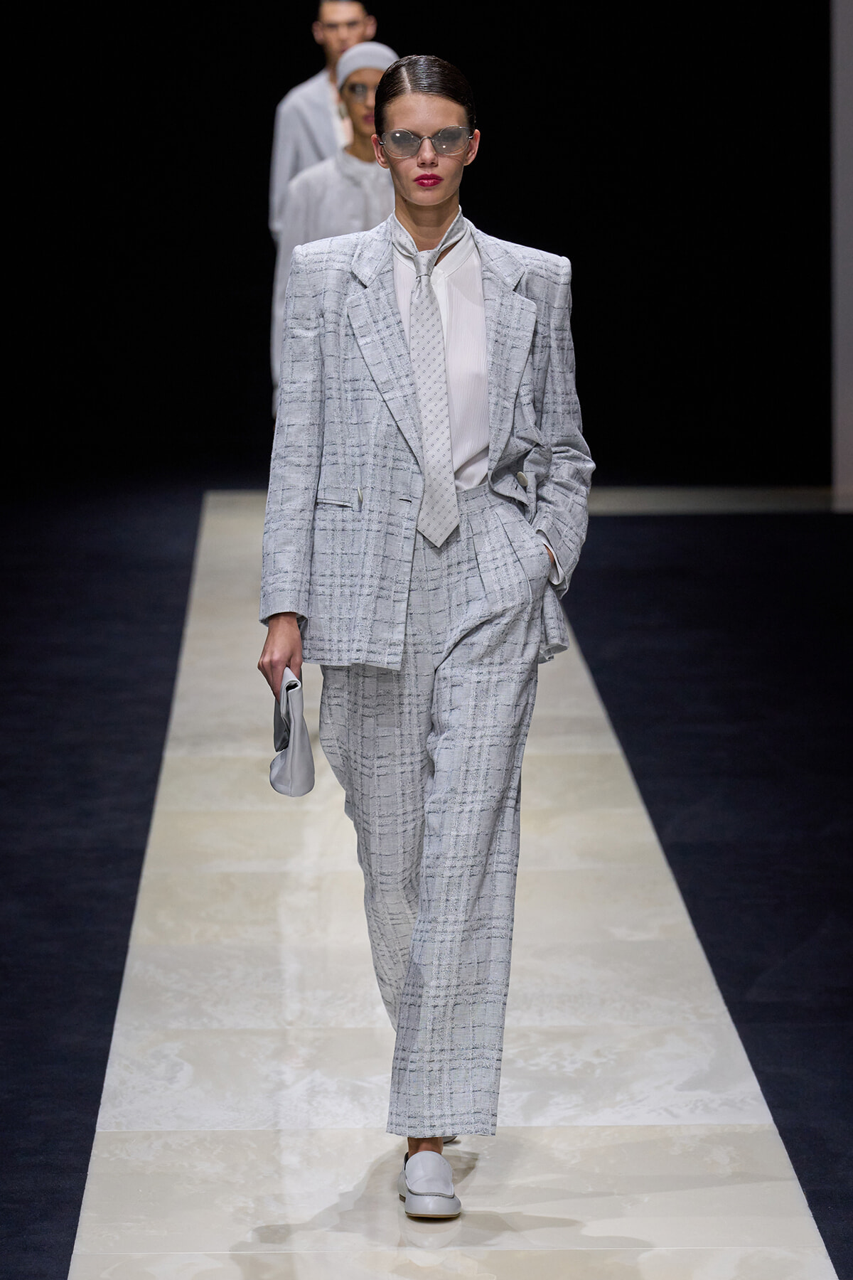 Model wearing a light gray plaid suit with oversized blazer and matching trousers, white shirt, pale tie, sunglasses, walking a fashion runway while holding a small light gray clutch.