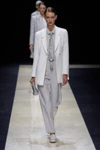 Model walking a fashion runway in an all-white suit with a light gray tie and matching pants, holding a small gray clutch.