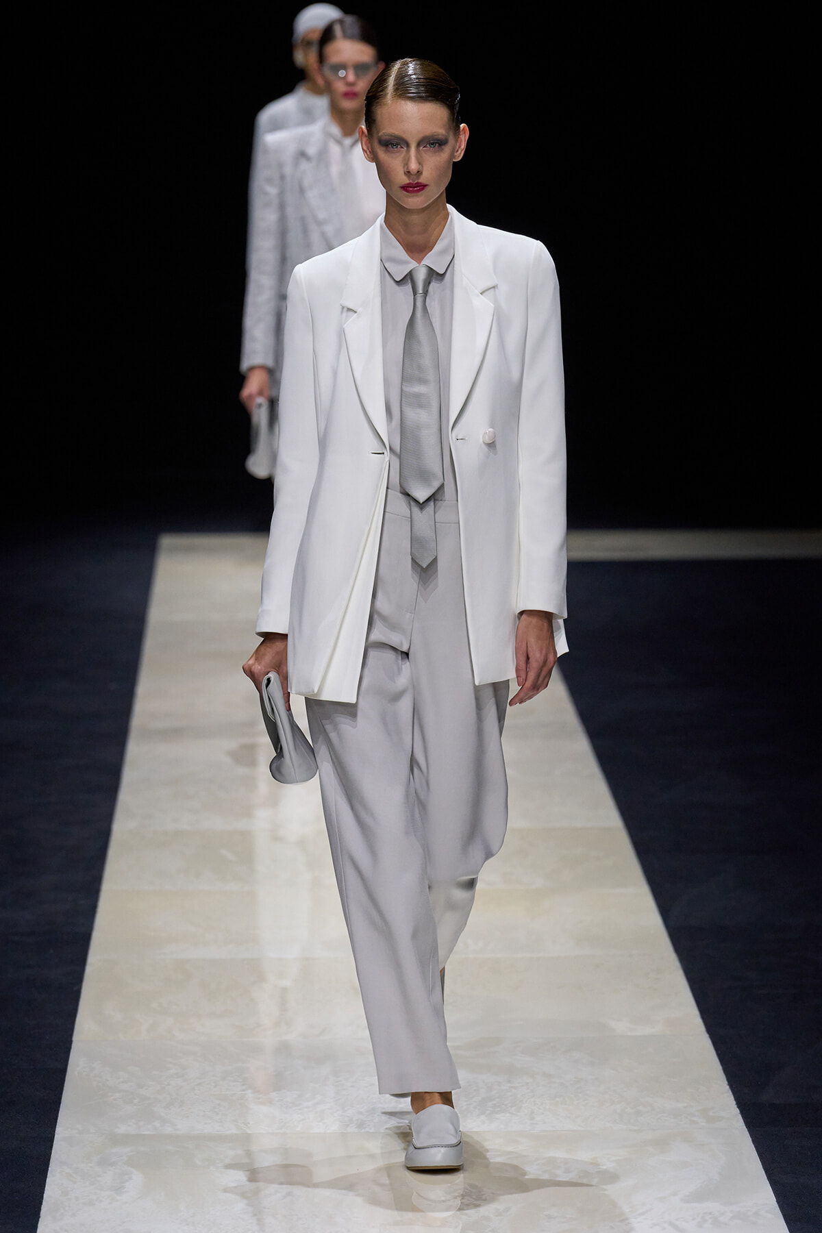 Model walking a fashion runway in an all-white suit with a light gray tie and matching pants, holding a small gray clutch.
