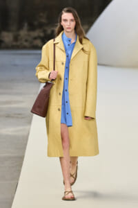 Model walking a fashion runway in a pale yellow textured coat over a blue shirt-dress, carrying a brown shoulder bag and wearing brown sandals.