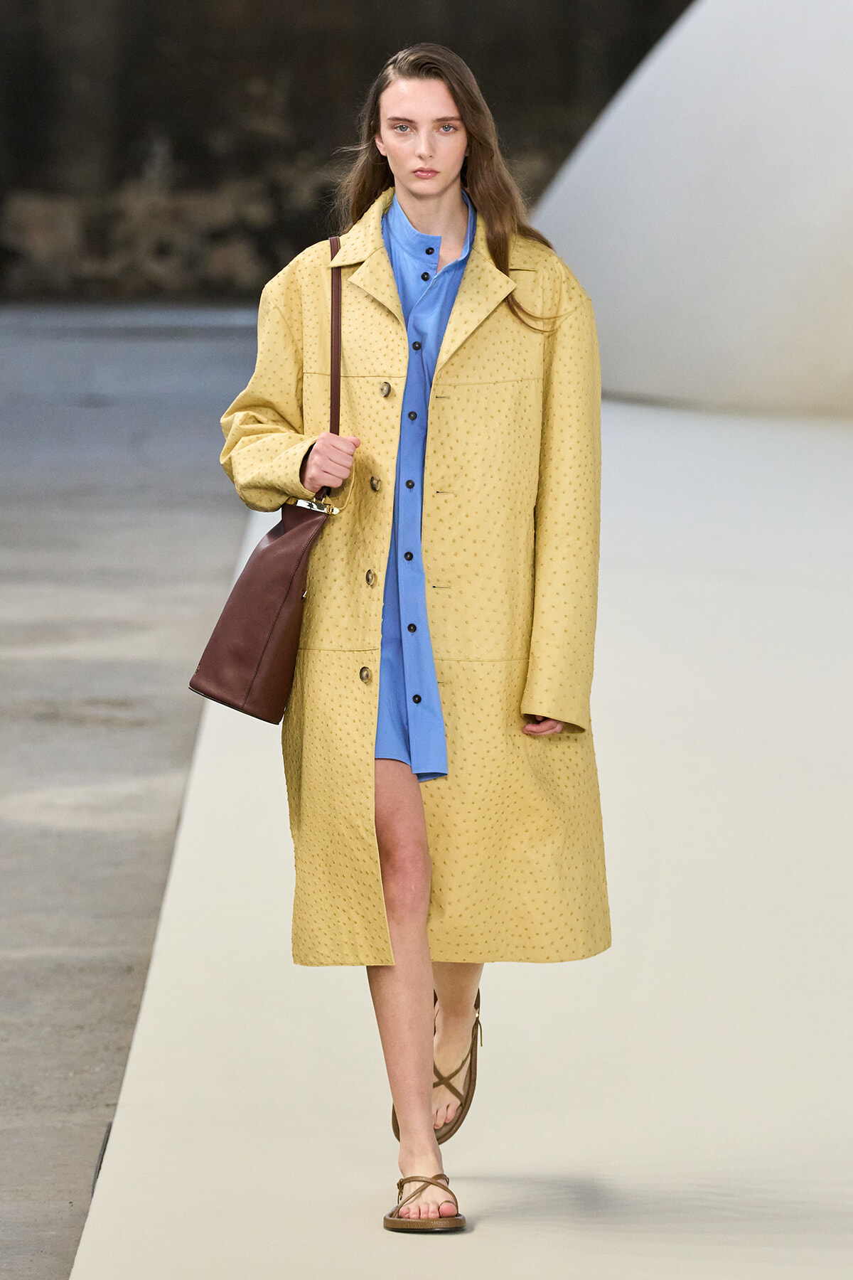 Model walking a fashion runway in a pale yellow textured coat over a blue shirt-dress, carrying a brown shoulder bag and wearing brown sandals.