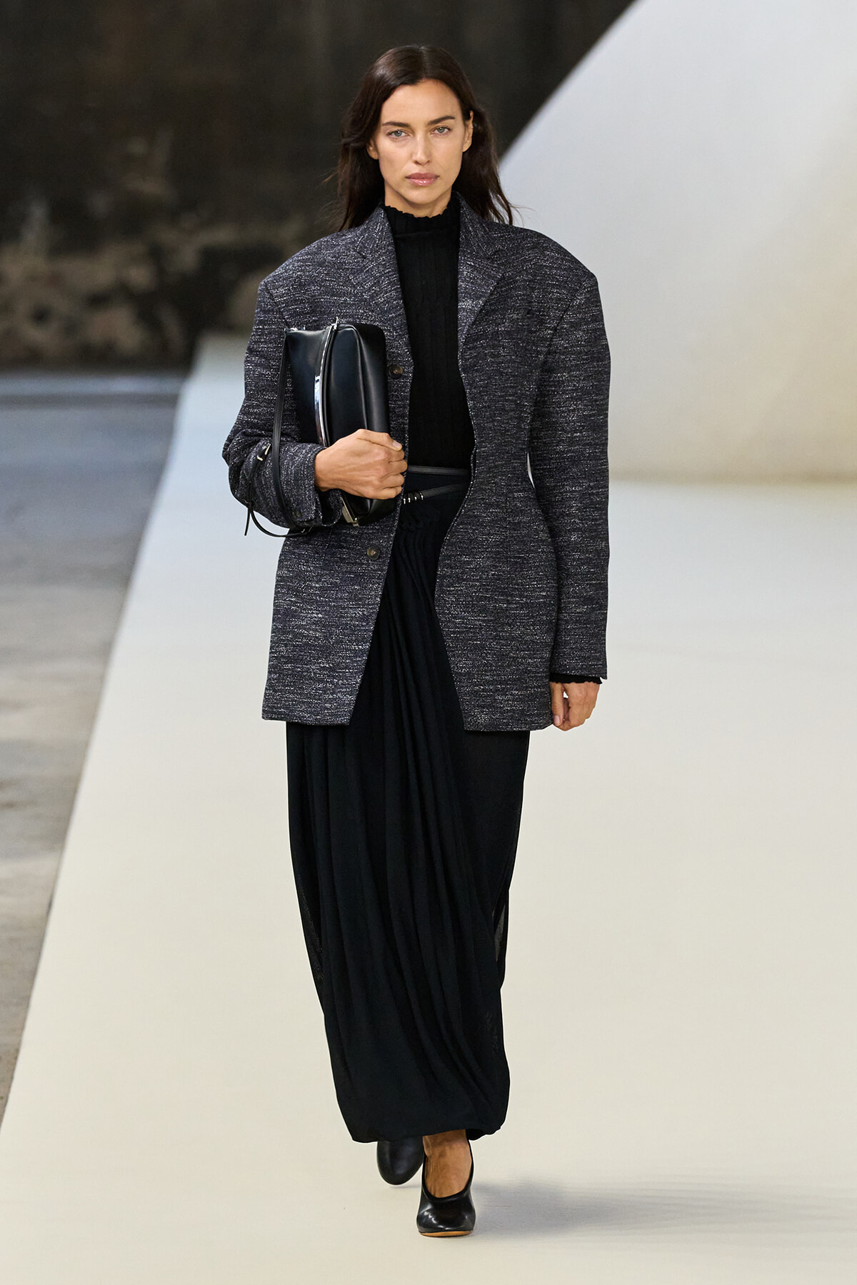 Woman walking a runway in a textured charcoal blazer over a black turtleneck and long draped skirt, carrying a black clutch.