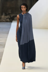 Model walks the runway in a navy sleeveless jumpsuit with a draped gray-to-navy cape cascading from the shoulder, during a fashion show