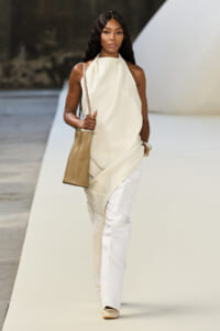 Model walking a runway in a cream draped halter top and white wide-leg pants, carrying a beige tote and gold jewelry.