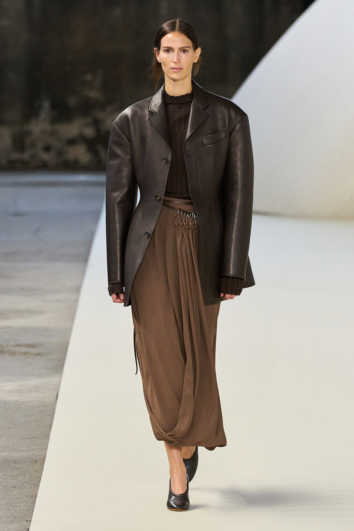 Model walking a runway in a oversized dark brown leather blazer, brown knit top, and draped tan skirt with black heels.
