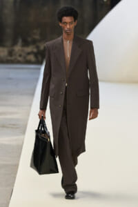 Male model walks a runway in a dark brown long blazer and matching trousers, carrying a black tote bag.