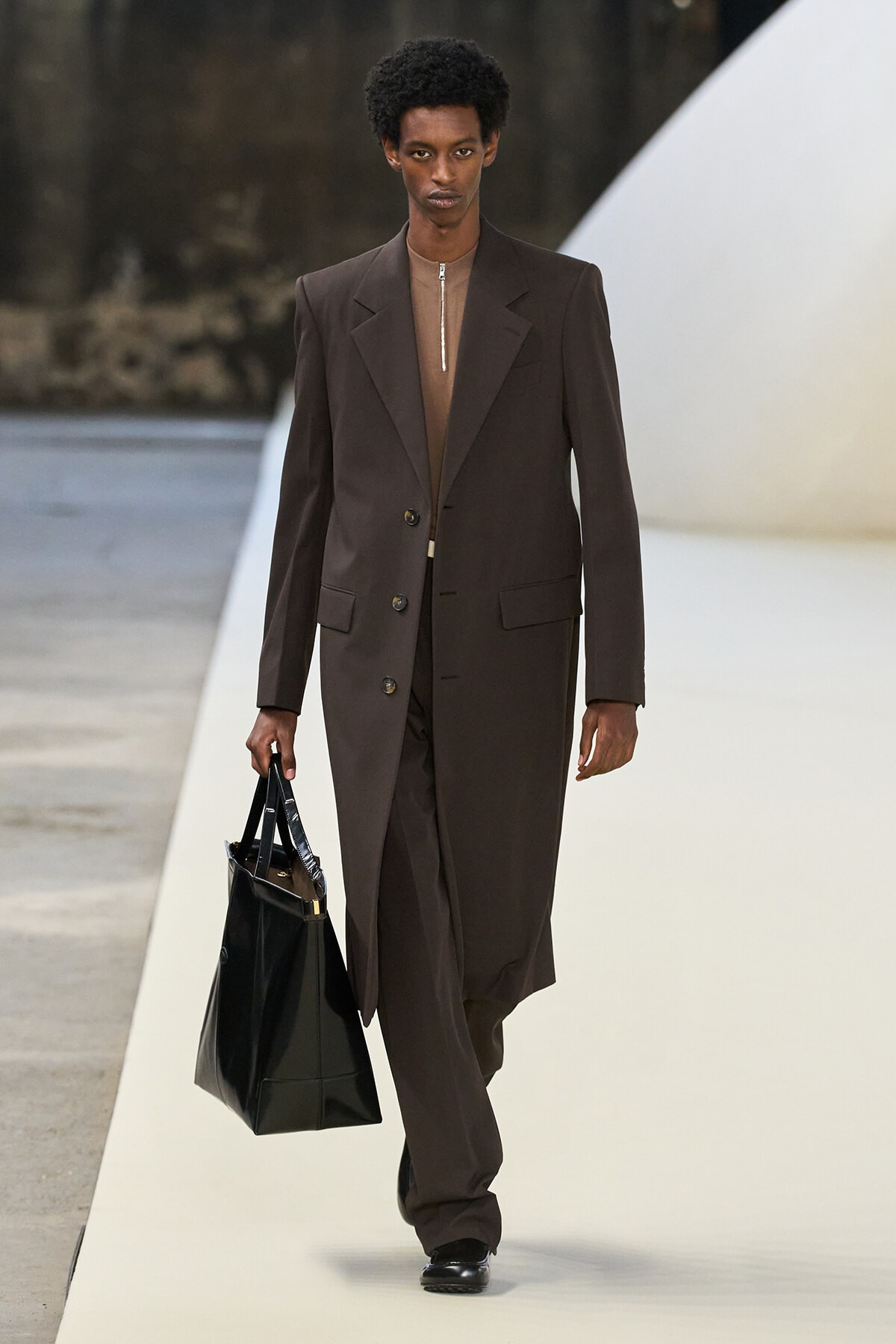 Male model walks a runway in a dark brown long blazer and matching trousers, carrying a black tote bag.