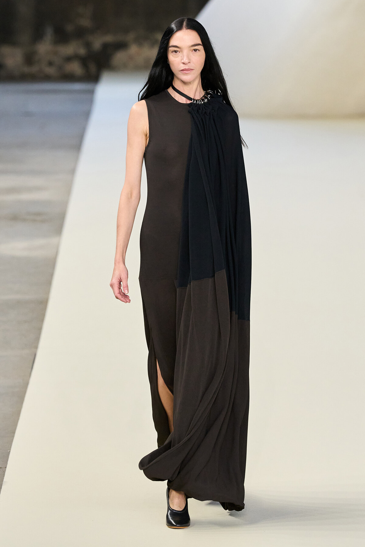 Model walking a fashion show wearing a sleeveless black dress with a long draped cape over one shoulder and black shoes.