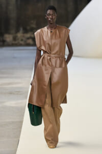 Runway model wears a tan leather sleeveless top with matching wide-leg belted pants, carrying a dark green crocodile-textured bag.