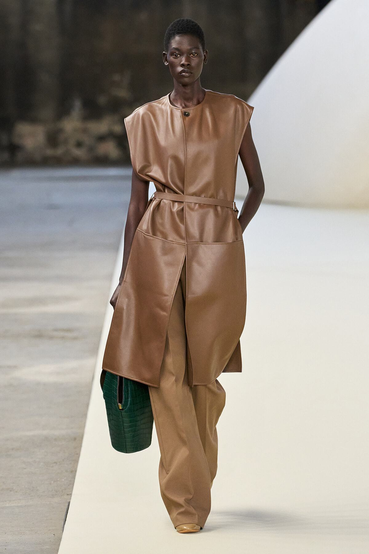 Runway model wears a tan leather sleeveless top with matching wide-leg belted pants, carrying a dark green crocodile-textured bag.