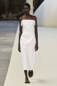 Model with dark skin wearing an off-the-shoulder white midi dress walking a fashion runway.