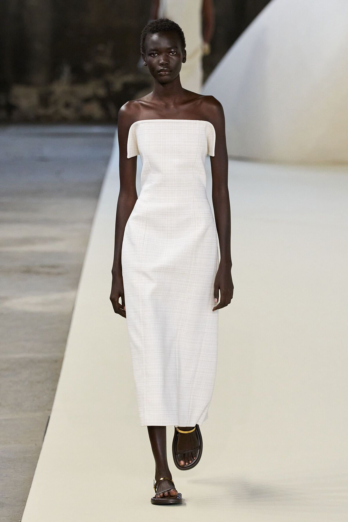 Model with dark skin wearing an off-the-shoulder white midi dress walking a fashion runway.