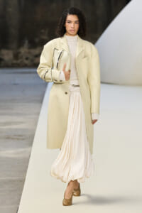 Runway look: oversized pale beige coat over a cream knit top and flowing ivory skirt, with a matching bag under the arm.