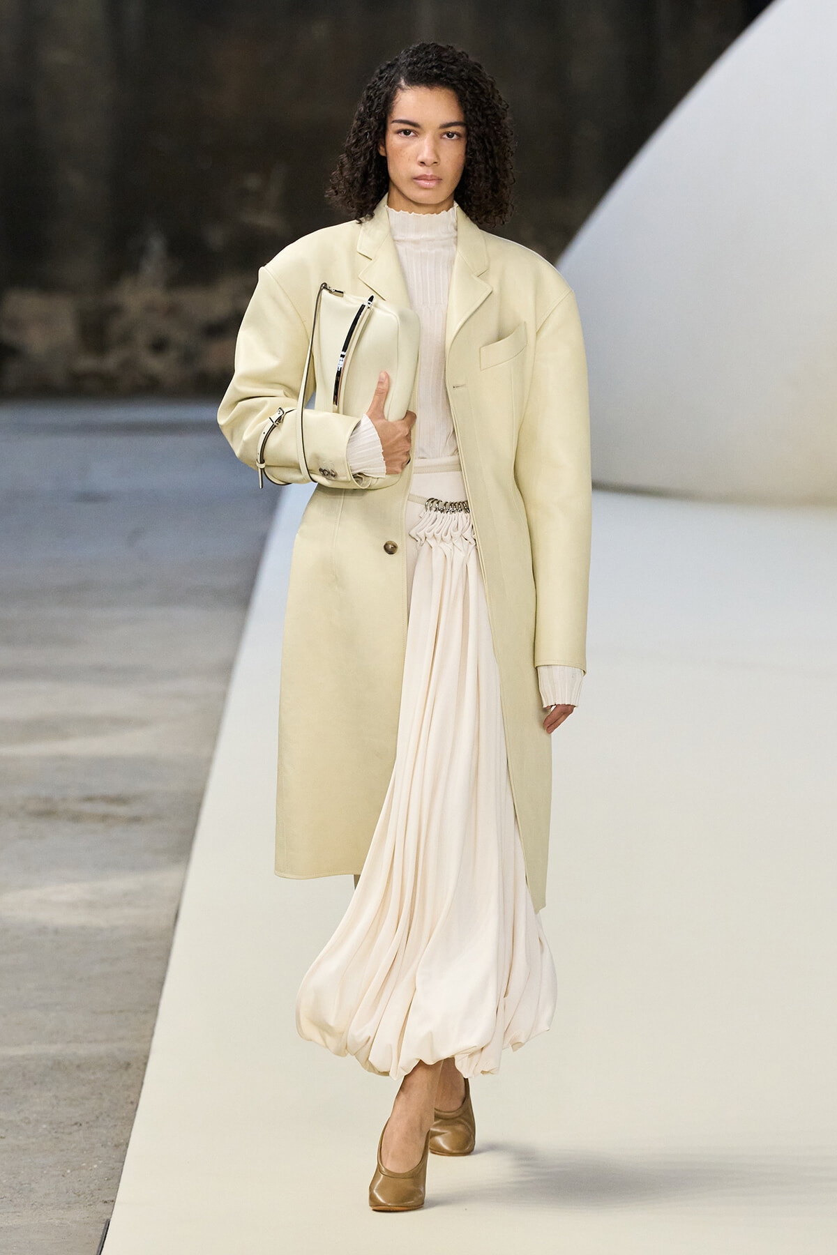 Runway look: oversized pale beige coat over a cream knit top and flowing ivory skirt, with a matching bag under the arm.
