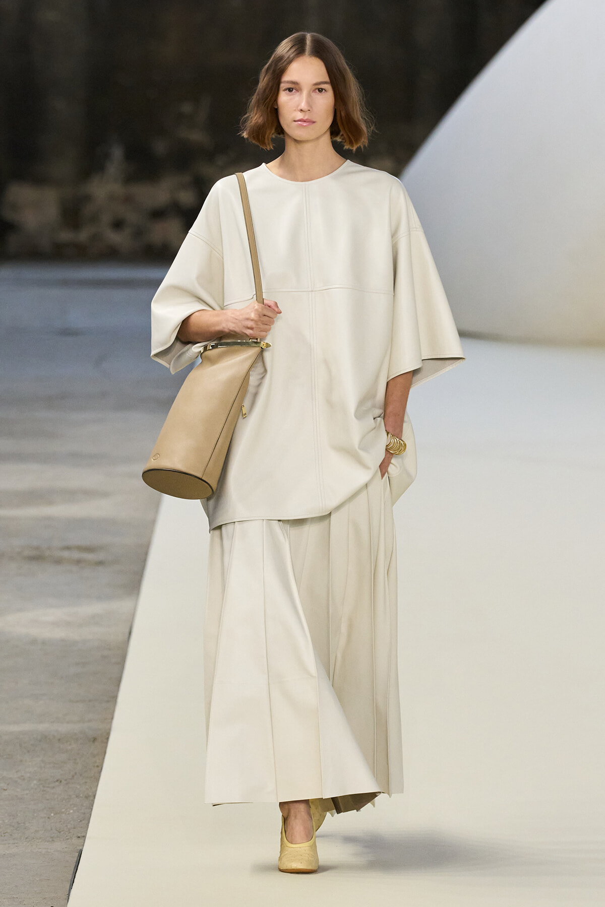 Model walking a runway in a cream oversized top with wide sleeves and matching wide-leg skirt, carrying a tan shoulder bag and wearing yellow shoes.