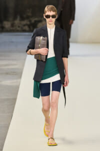 Model walking a runway in a dark blazer over a white top, color‑blocked green and navy shorts/skirt, yellow sandals, and oversized sunglasses, carrying a brown clutch.