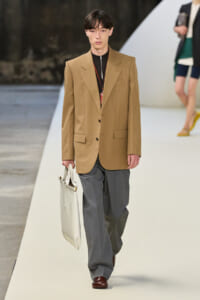 Male model on a runway wearing a tan blazer, dark top, gray wide-leg pants, dark brown shoes, and carrying a white tote bag.