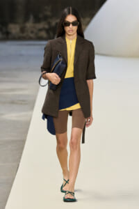 Model walks a runway in a brown blazer over a yellow top with a blue color-block panel, brown tights, green strappy sandals, dark sunglasses, and a navy clutch.