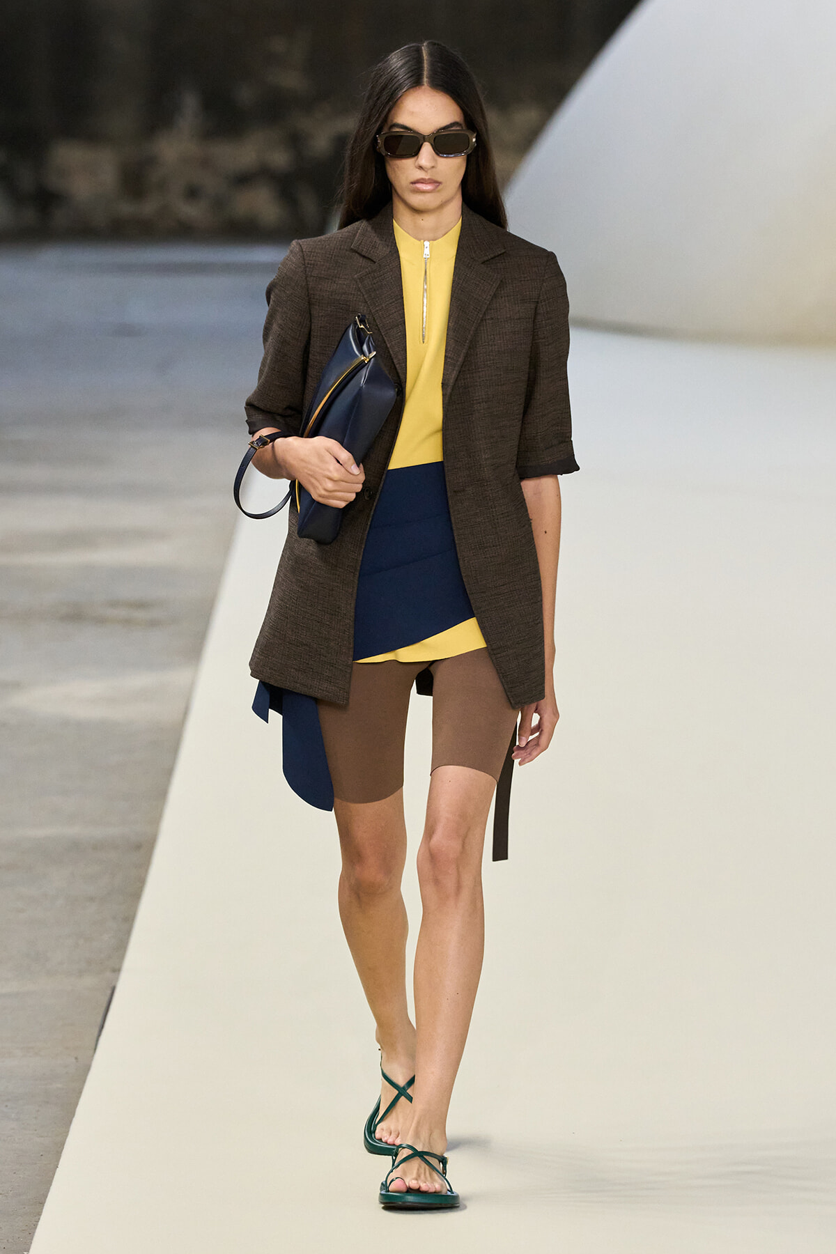 Model walks a runway in a brown blazer over a yellow top with a blue color-block panel, brown tights, green strappy sandals, dark sunglasses, and a navy clutch.