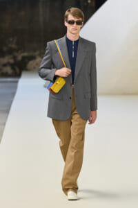 Male model on a runway wearing a gray blazer, navy top, tan trousers, white shoes, sunglasses, and a colorful crossbody bag.