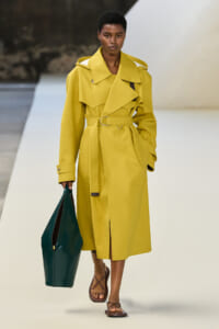 Model walking runway in a bright yellow trench coat with belt, carrying a dark green tote and wearing brown sandals.