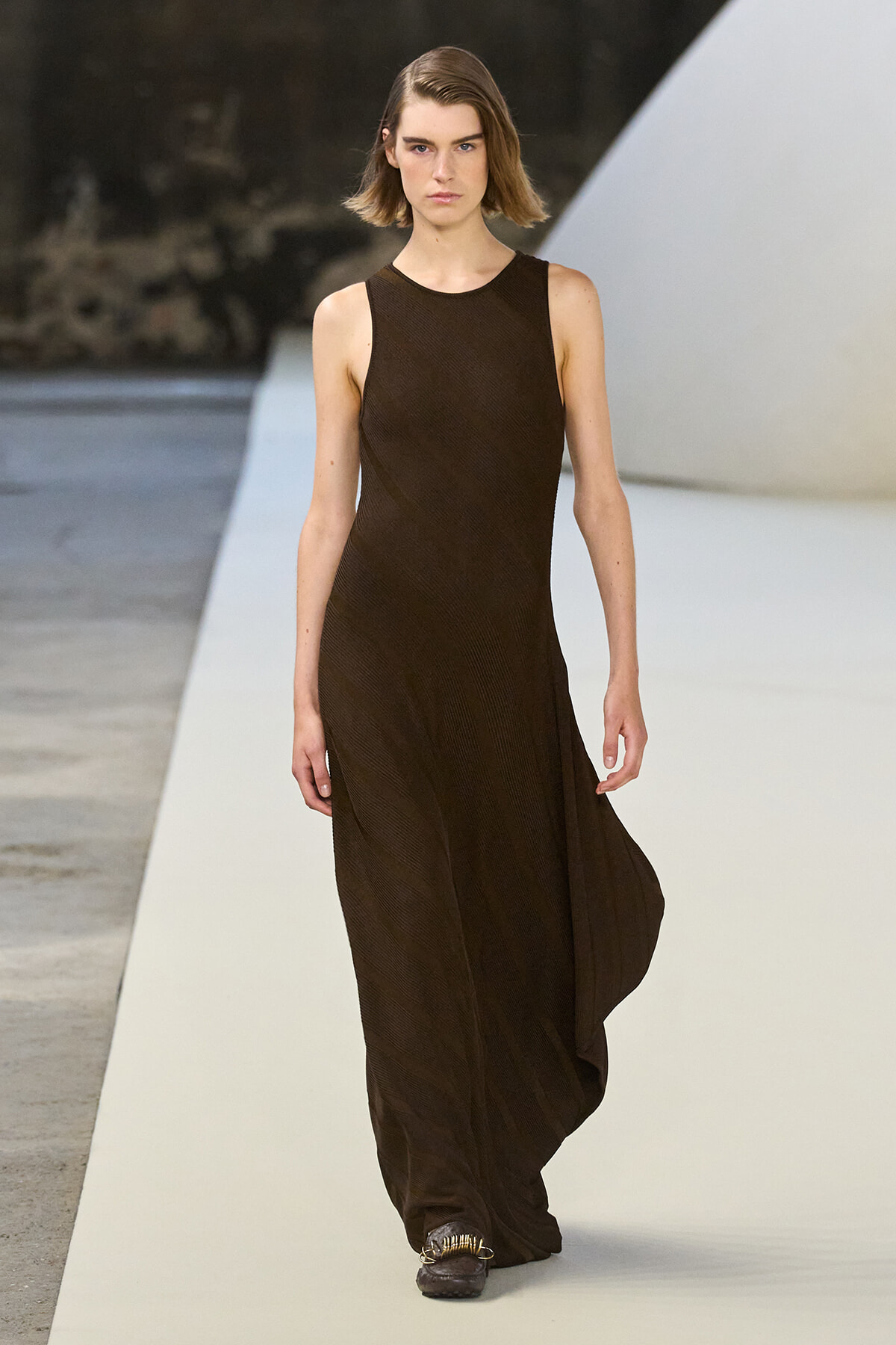 Model walking a fashion runway in a sleeveless dark brown maxi dress.