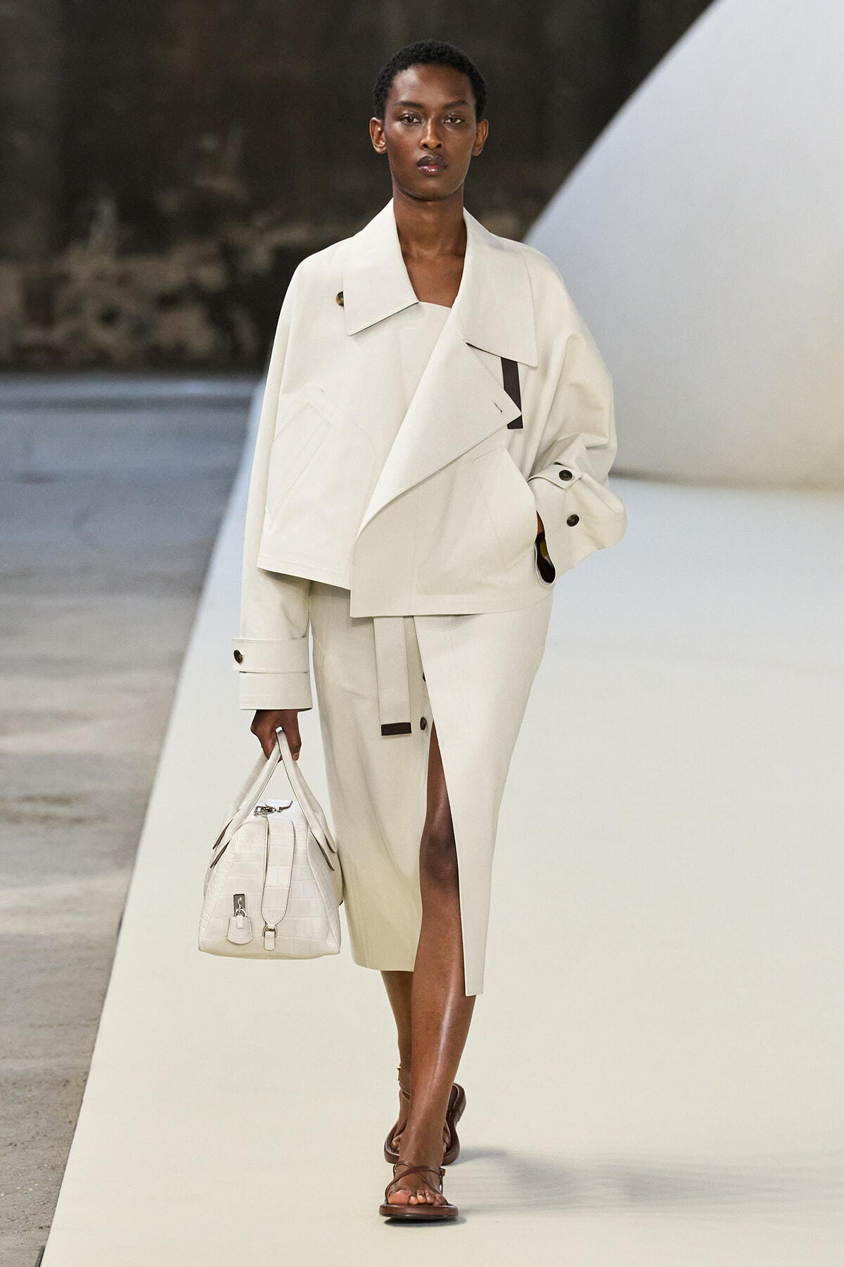 Model walking a runway in a large off-white trench coat with a matching handbag, brown sandals, and a short natural hairstyle.