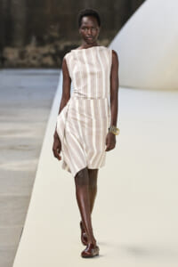 Model walks a runway in a sleeveless beige-and-white striped dress with a wrapped skirt and gold bangles.
