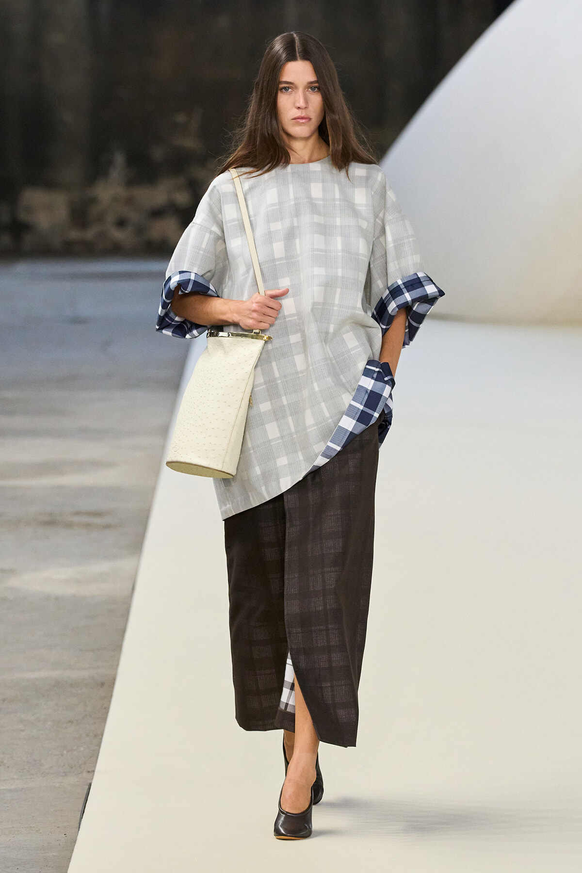 Runway model in a light gray plaid oversized top with blue plaid cuffs, a dark plaid skirt, black heels, and a cream bucket bag.