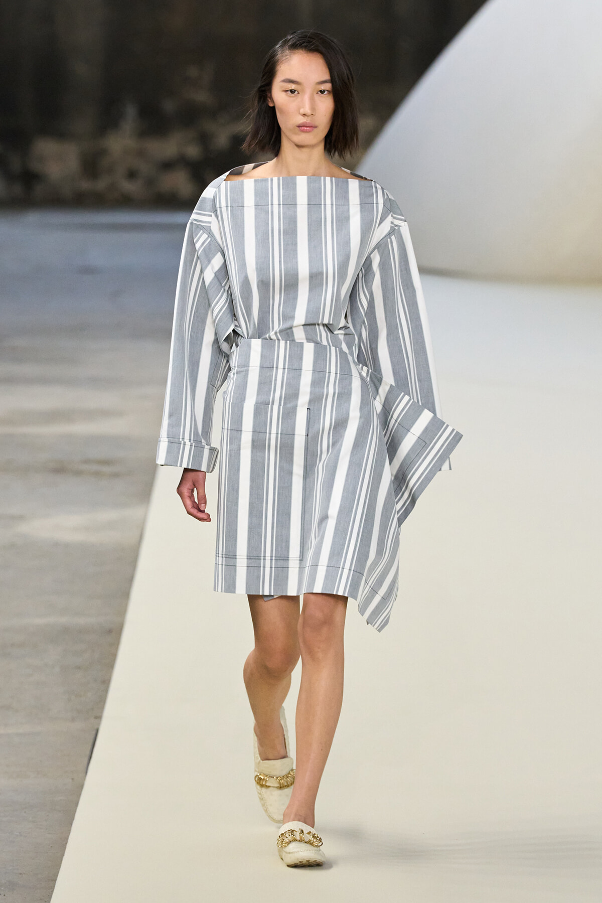Model walking the runway in a loose gray-and-white vertical striped dress with oversized sleeves and a belted waist.