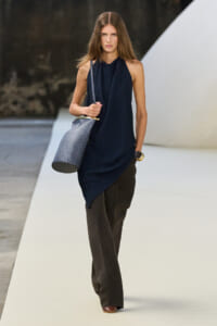 Runway model walks in a navy draped sleeveless top with dark wide-leg pants, carrying a gray textured bucket bag.