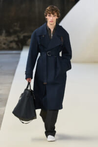 Male model walking a runway in a dark navy trench coat with a belt, carrying a black structured bag and wearing white sneakers