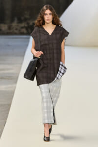 Model walking the runway in a loose dark plaid V-neck tunic paired with light gray plaid pants, carrying a black shoulder bag.