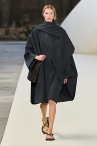 Model walks a runway in a dark oversized cape coat, carrying a black shoulder bag and wearing strappy sandals.