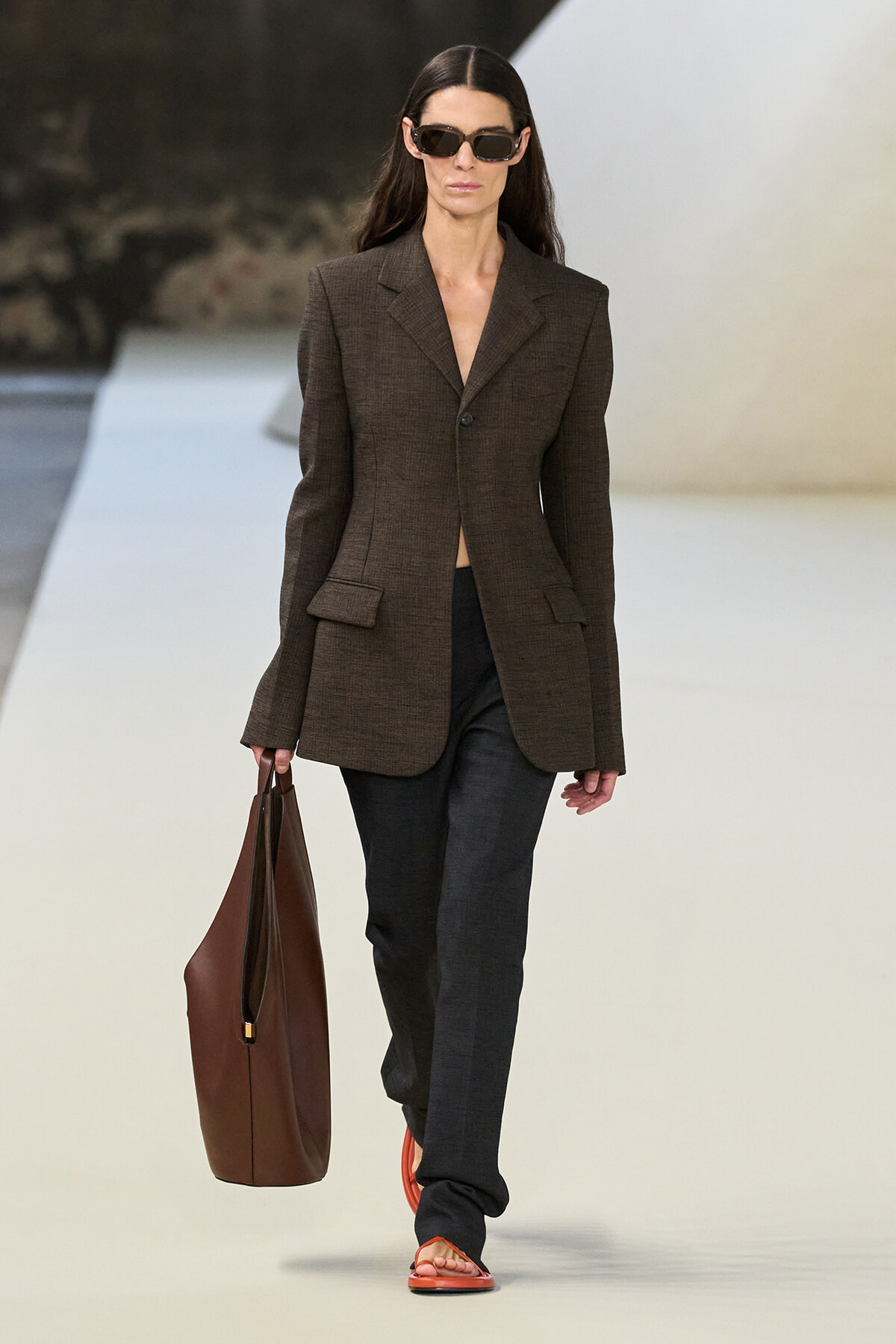 Model walking a runway in a brown blazer, dark trousers, sunglasses, and a large brown tote bag.