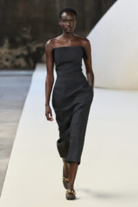 Model walking the runway in a strapless charcoal midi dress and brown sandals with gold chains.