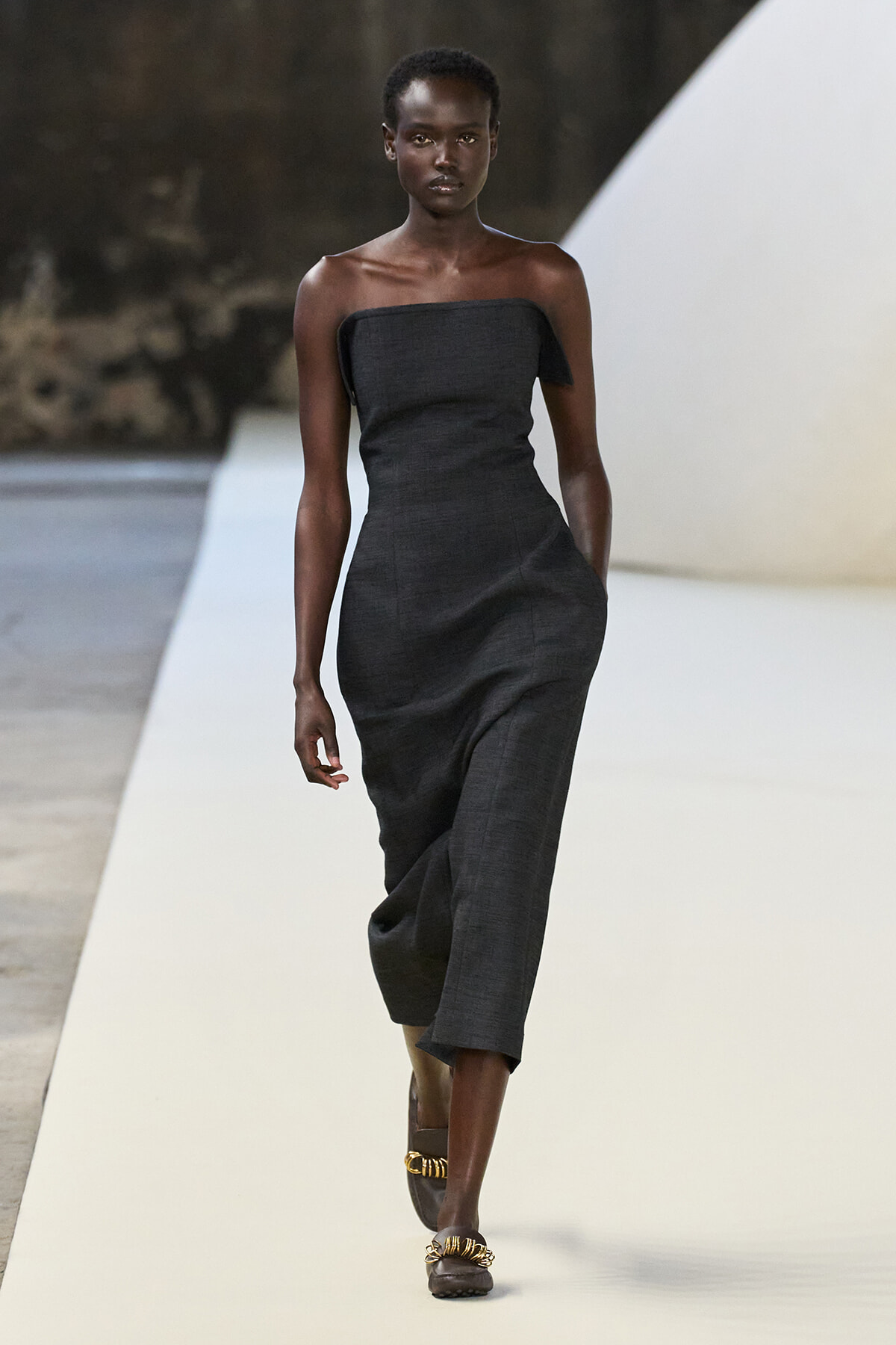 Model walking the runway in a strapless charcoal midi dress and brown sandals with gold chains.
