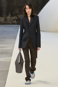 Female model walks a runway in a dark tailored blazer and brown trousers, carrying a gray handbag and wearing bright blue sandals.