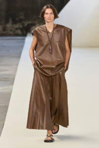 Model walking a fashion runway in a brown sleeveless leather top and matching wide-leg skirt, with a pendant necklace and sandals.