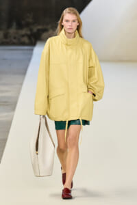 Runway model wearing an oversized pale yellow jacket with green shorts, burgundy loafers, and a white tote bag.