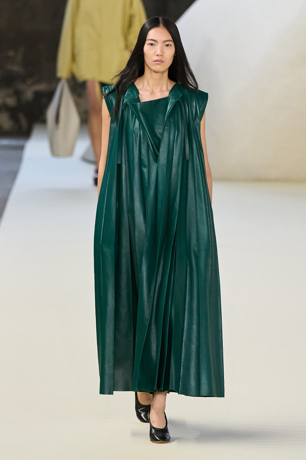 Model walking a fashion runway in a long, emerald green sleeveless dress with structured pleats and a glossy leather-like finish.